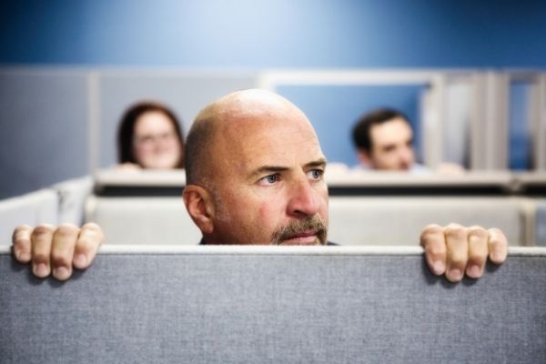 man in a work cubible peering over the top of the partition