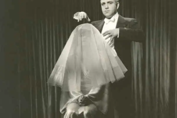 woman wearing a veil and sittting on a chair on a stage with a stage hypnotist standing behind her looking at the audience